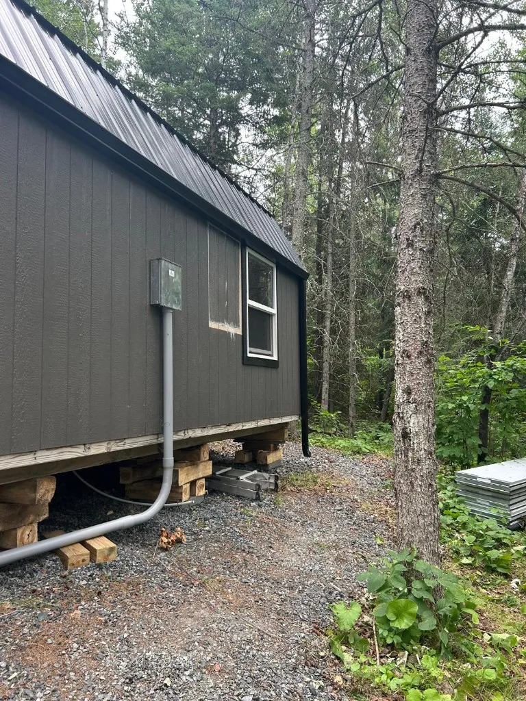 Black eavestrough and downspout on a dark board-and-batten cabin with a black metal roof, set in a spruce forest near Kenora