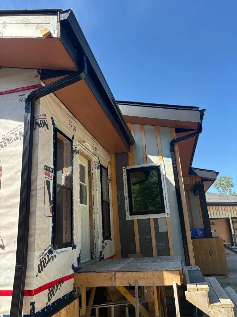 Black seamless eavestrough and downspouts installed on a new build still wrapped in house wrap, with cedar-lined soffit overhangs