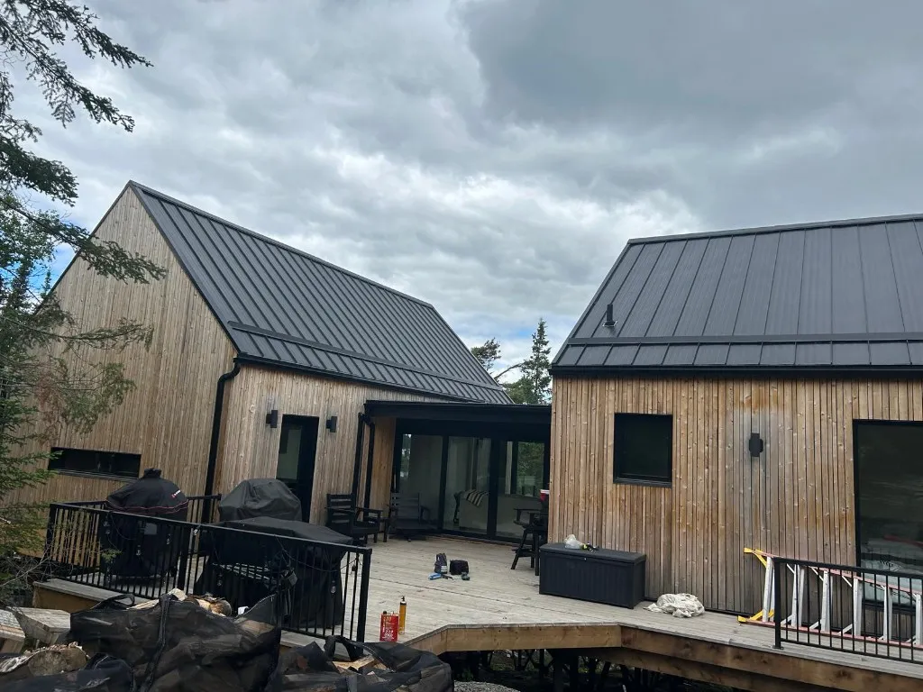 Two-gable cottage clad in vertical cedar with a black standing-seam metal roof, black seamless eavestrough and matching downspouts above a large deck