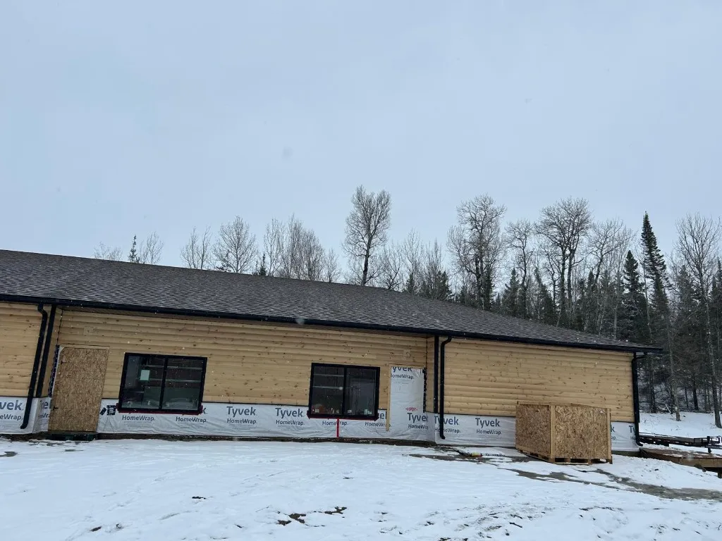Single-storey building with asphalt shingles and black eavestrough installed over snow-covered ground, walls still partly wrapped