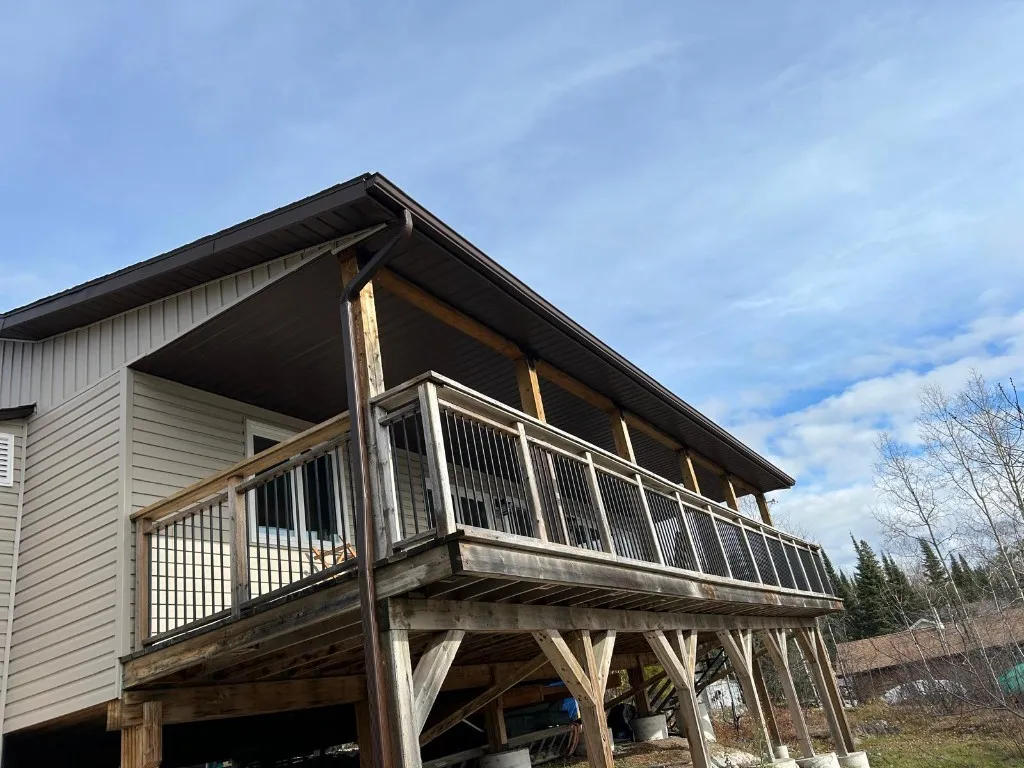 Brown eavestrough and downspout following the roofline of a covered elevated deck on a beige vinyl-sided home