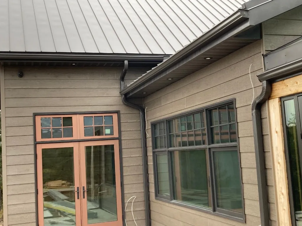 Close-up of dark bronze eavestrough, downspout and pot-lit soffit above the windows of a taupe lap-sided home with a dark metal roof