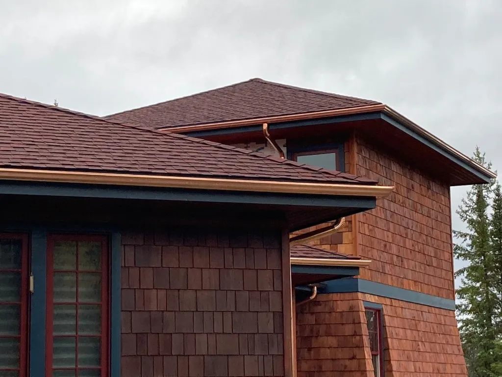 Copper eavestrough and copper downspout on a cedar-shingle home with a red-brown shingled roof and navy trim
