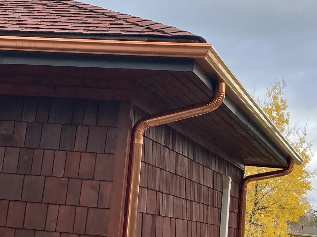 Close-up of a copper eavestrough and copper downspout with crimped elbows running down cedar shake siding, autumn foliage behind