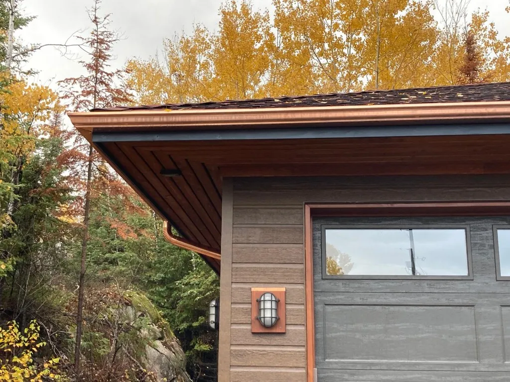 Copper eavestrough, copper downspout and copper light backplate on a garage with stained wood soffit, framed by golden birches
